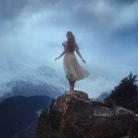 Девушка в платье со спины, которая стоит на камне на фоне гор.