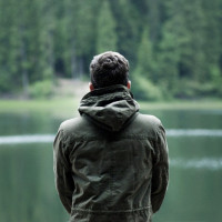 Стоящий у берега озера мужчина в куртке со спины