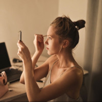 Девушка с зеркальцем в руках красит брови за своим рабочим столом