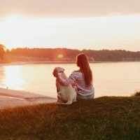 Девушка с собакой смотрят на восход солнца неподалёку от воды