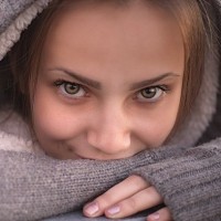 Девушка в тёплом капюшоне мило улыбается, прикрыв губы рукой.